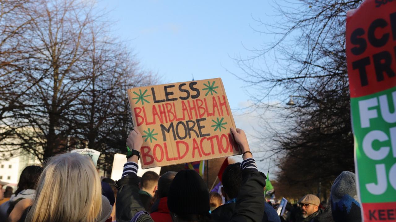 Protest walking in between avenue of trees with a placard centre shot reading 'less bla blah blah, more bold action'