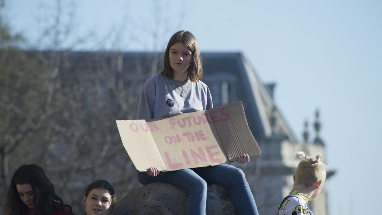 Young Climate protestor sits holding cardboard on which is written 'Our future is on the line'.