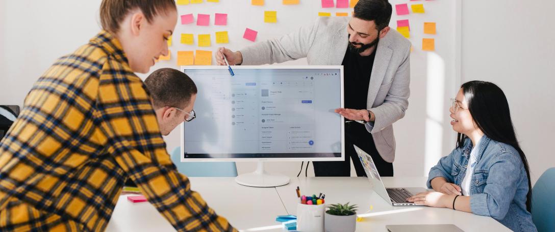 Photo of people working around a table with someone pointing at a presentation. Photo by Jason Goodman on Unsplash