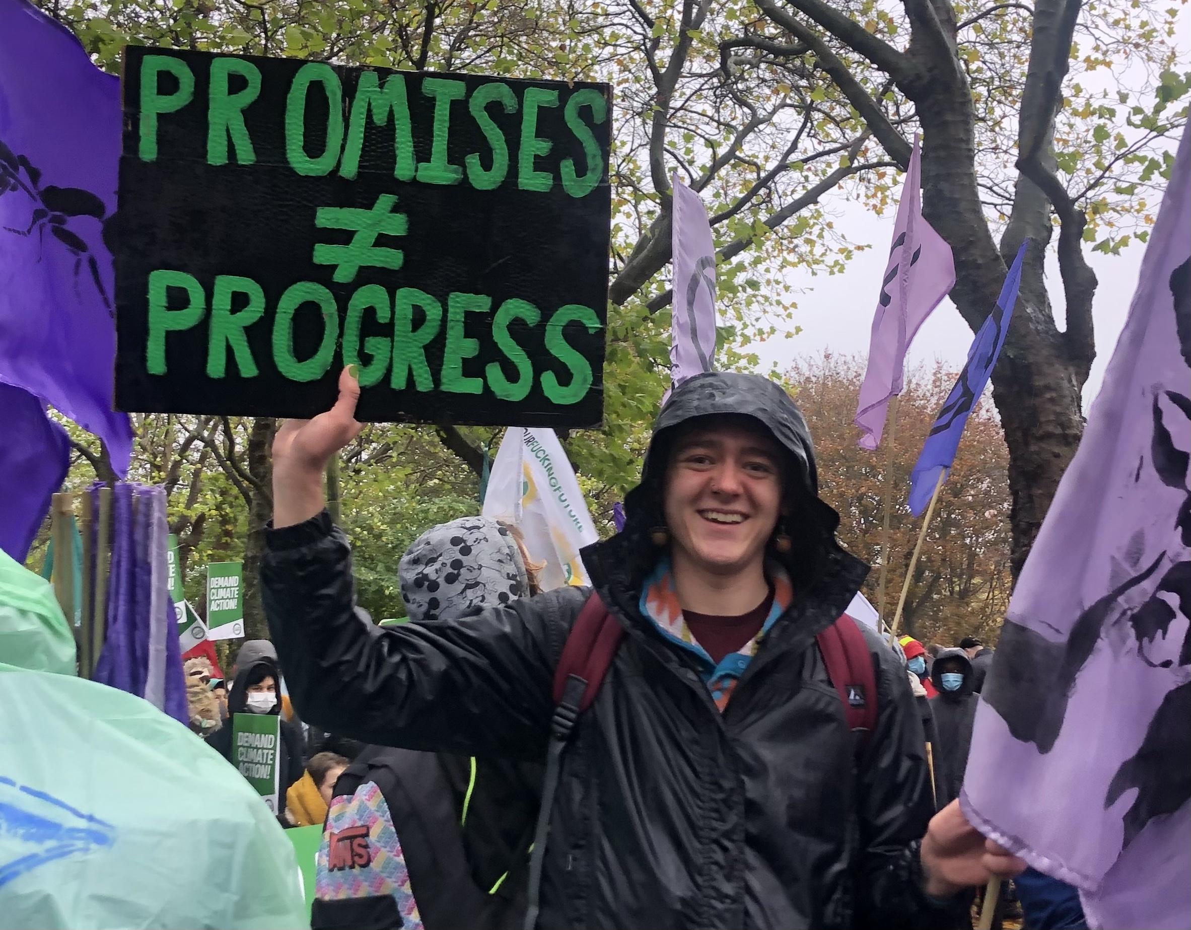 Nick Koeing, in a wet rain jacket at a protest, holding a placard with a black background and writing in green reading 'promises do not equal progress'