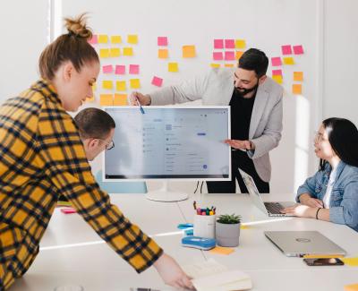Photo of people working around a table with someone pointing at a presentation. Photo by Jason Goodman on Unsplash